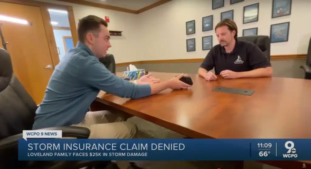 Two men sit at a large wooden table in an office setting, discussing a storm insurance claim, as indicated by the WCPO 9 News banner at the bottom of the screen which reads 'STORM INSURANCE CLAIM DENIED, LOVELAND FAMILY FACES $25K IN STORM DAMAGE.'